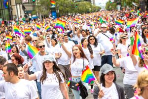 2015 Pride Parade in San Francisco featuring the sponsorship of Apple. All of the people marching are holding up small rainbow pride flags and wearing white shirts with a minimal Apple logo in the center of them. Some people have their mouths open as if they're chanting or singing, others are just walking. The crowd is massive and defocuses into a sea of rainbow flags.