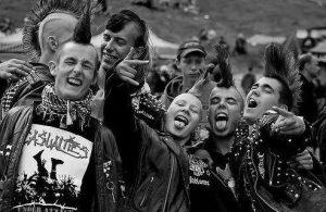 Group of people in the punk subculture posing for a picture. They are all sporting mohawks and look to be happy. They are wearing leather jackets with studs, and one of them has a lip piercing. The image is in black and white and one person is throwing up a "rock on" sign and another a "peace" sign with their hands. 