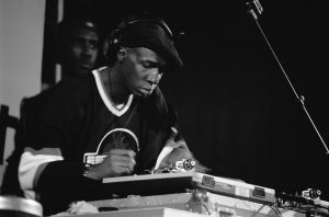 A black and white photograph of Grandmaster Flash, one of the original DJ's, scratches a disc on stage at a performance