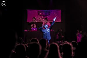 Hopsin stands on stage performing at a small, underground hip-hop club. Behind him stand sound mixers and the rest of his group.