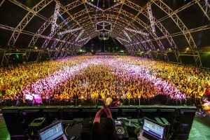 Thousands of people cheer and dance for DJ Bassnectar at Coachella. The image is from the perspective behind the DJ, and shows Bassnectar with his hands raised about his computer and soundboard as he dances and looks into a very large crowd in Coachella's (notable) Sahara tent where most EDM acts perform. The audience is showered in yellow and pink light below the tent's dark atrium. The crowd reaches far back in the tent and possibly beyond.