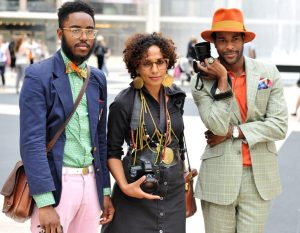 3 people stand with vintage but clean-cut suit outfits on holding cameras. They are wearing bright colors or eccentric jewelry. 