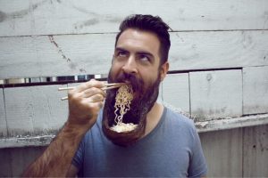 Graphic shows a man whose beard has been matted into a bowl shape and is shoveling ramen noodles from it into his mouth using chopsticks. He looks up in a way that could mean he sees his actions as ironic.