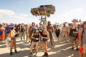 People at Burning Man pose in front of a spray-painted car. They surrounded by people, all dressed differently. The two people in the foreground are dressed lightly for the warm weather. They are wearing shorts and tank tops. One person is sporting a floppy blue hat. They are both smiling.