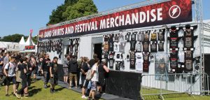 Merchandise booth at a concert showing many t-shirts on a wall, with people gathering around the fenced off area with the seller inside. The banner at the top of the booth says "Band shirts and festival merchandise"
