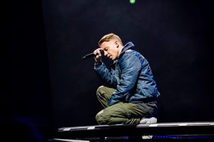 Macklemore kneels with his eyes closed and a microphone leaned against his head during a song.