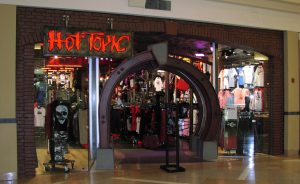 The front of the retail store Hot Topic. Hot Topic is written in kind of creepy red text and the entrance to the store is a dark archway. In the indow is a show with a skull on it, and the inside looks dark and boasting with merchandise. 
